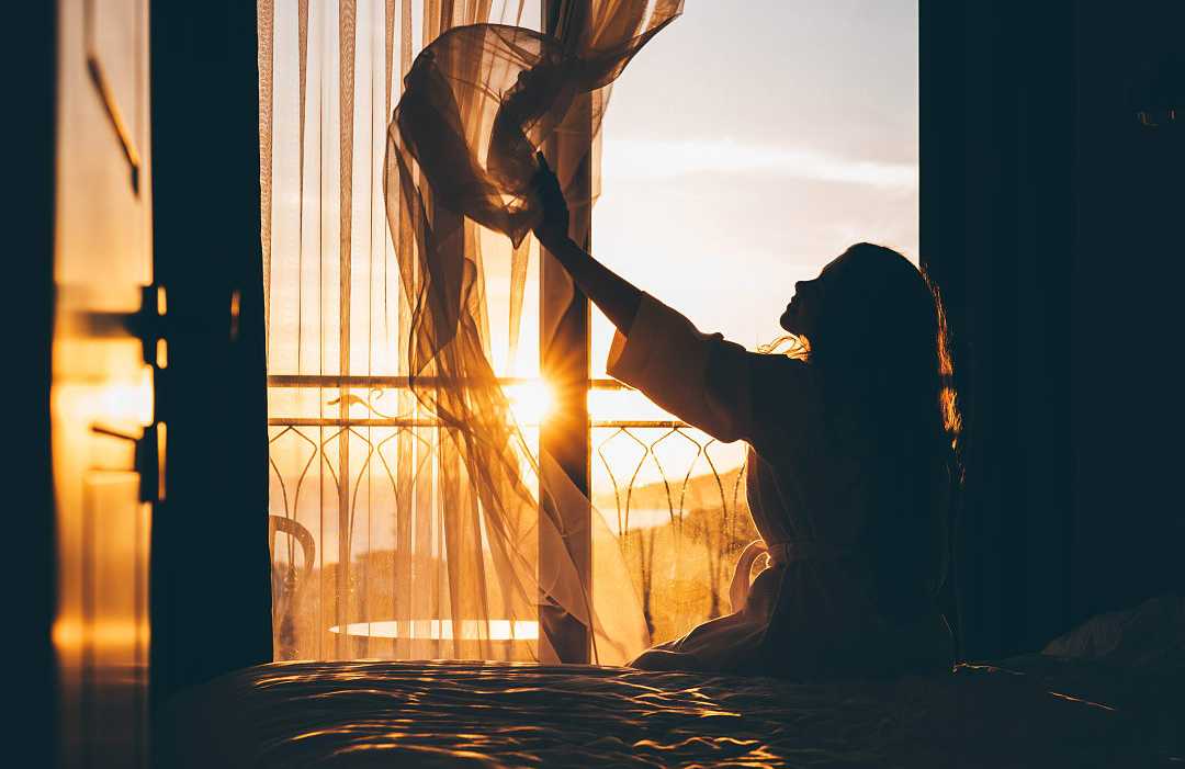 Woman sitting on a bed and opening curtains as the morning sunlight streams into a hotel room. Woman sitting on a bed and opening curtains as the morning sunlight streams into a hotel room.