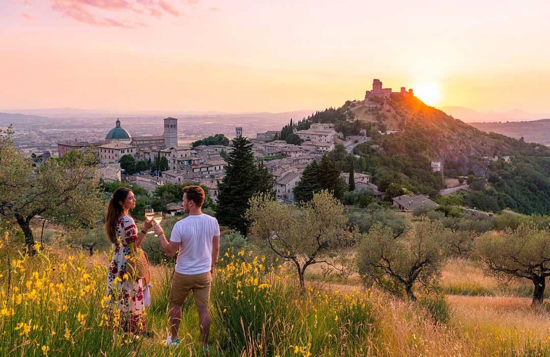Couple toasting with wine at sunset overlooking a picturesque medieval town surrounded by olive trees and rolling hills