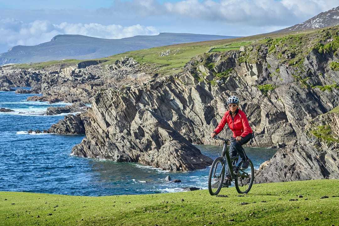 Senior cyclist biking along scenic Irish coastal cliffs with blue ocean waves and rugged landscapes