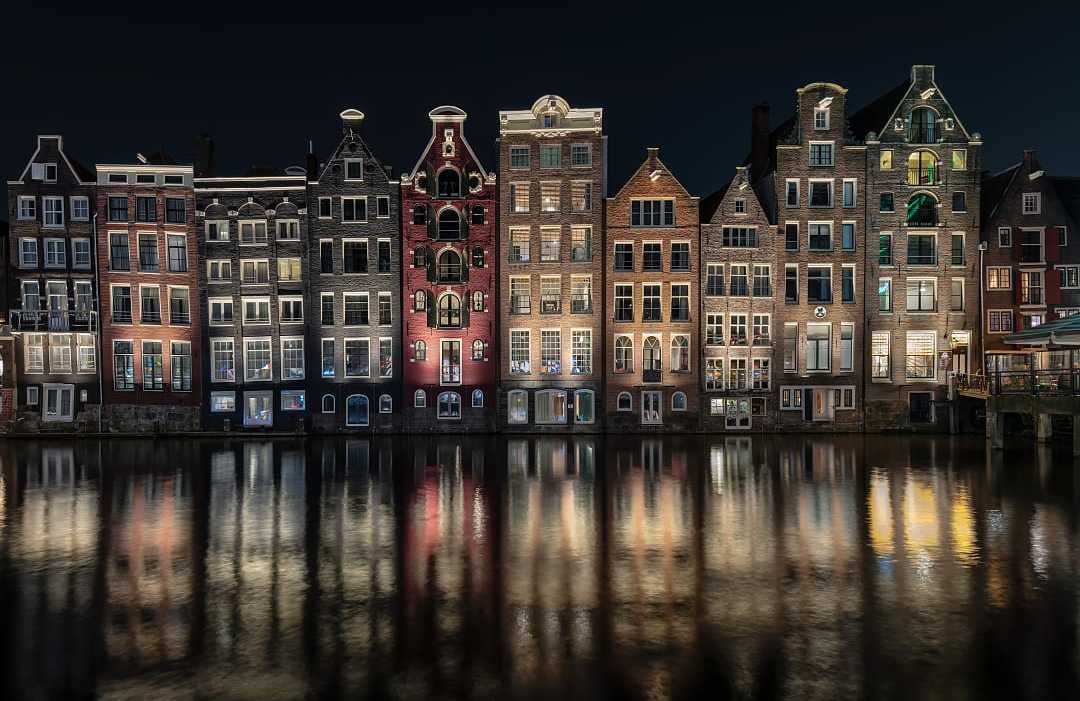 Illuminated traditional Dutch gabled houses reflecting in an Amsterdam canal at night.