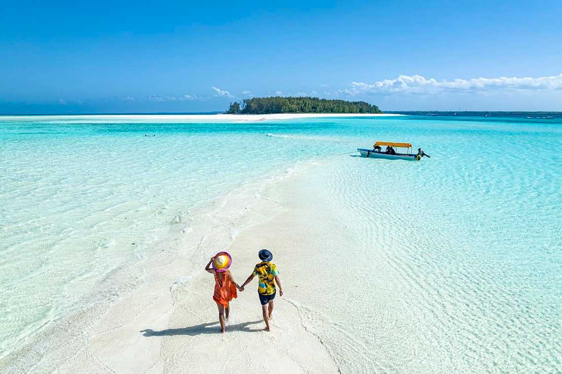 Couple walks on sandbank in clear turquoise water toward island in Zanzibar, Tanzania.