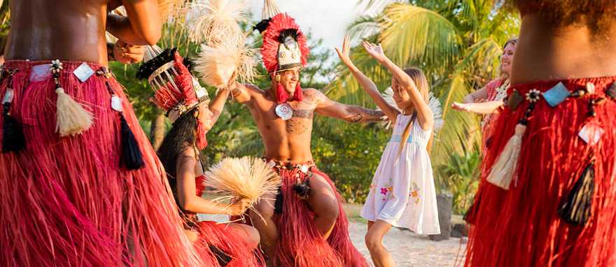Photo courtesy of Tahiti Tourisme Grégoire Le Bacon Young girl and her mother with Tahitian men and women in traditional attire performing a cultural dance surrounded by lush greenery and a serene ocean backdrop in Tahiti