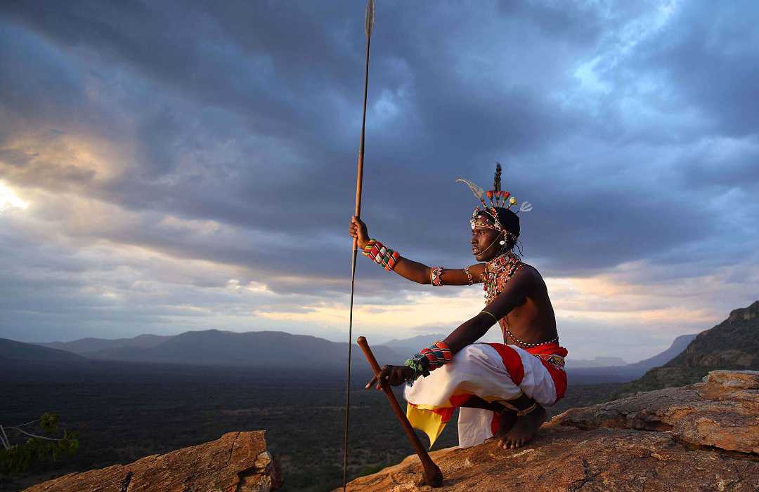 A traditional warrior sitting on a rocky hill at sunset, holding a spear and overlooking a vast wilderness landscape.